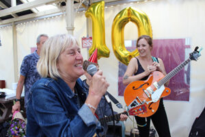 A woman holding a microphone and smiling. Behind her, there are balloons in the shape of a number 10 and a woman holding a guitar